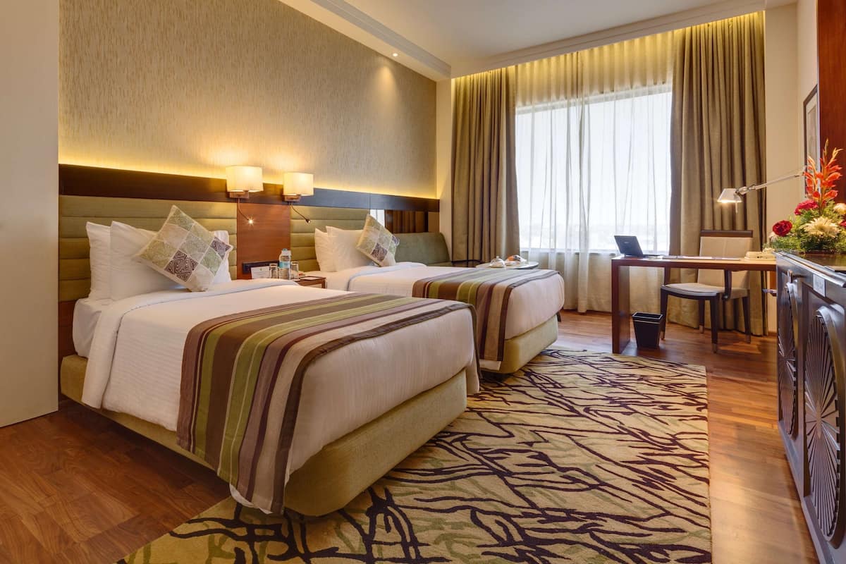 A hotel room with two beds, a desk, a chair, a lamp, and a large window with curtains.