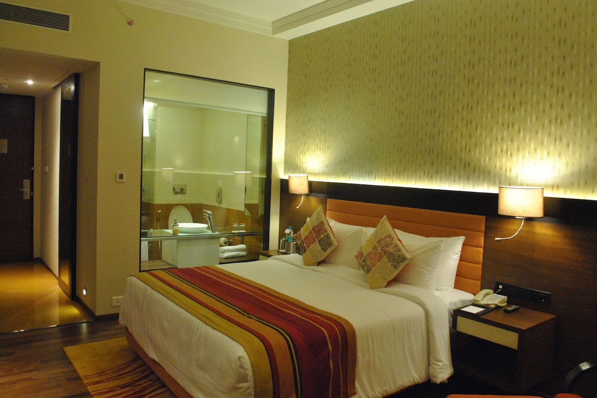 A hotel room with a large bed, a nightstand with a lamp, a mirror, and a bathroom visible through an open door.
