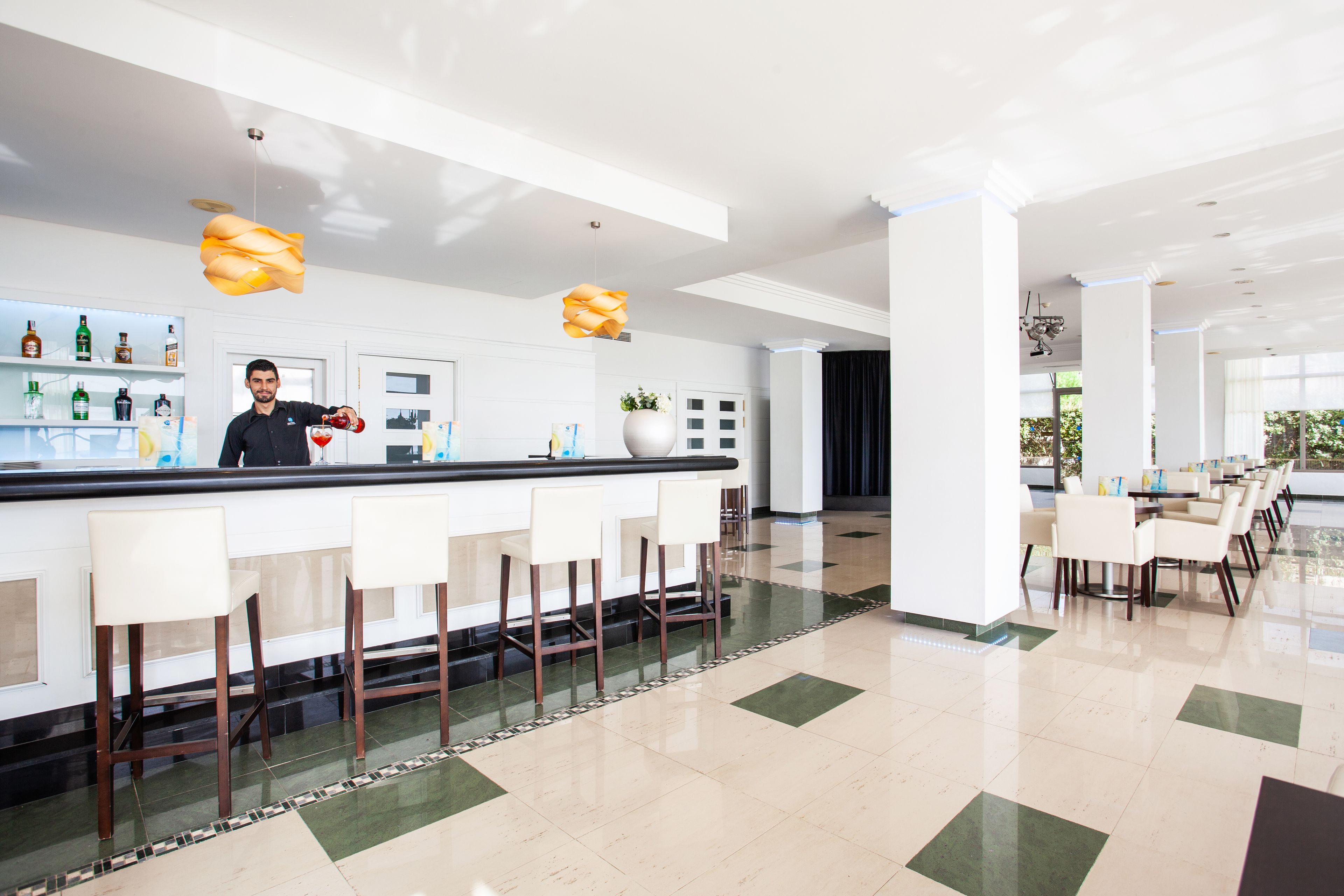 Un espace bar moderne avec un barman, des tabourets de bar et un comptoir où sont exposées différentes bouteilles.