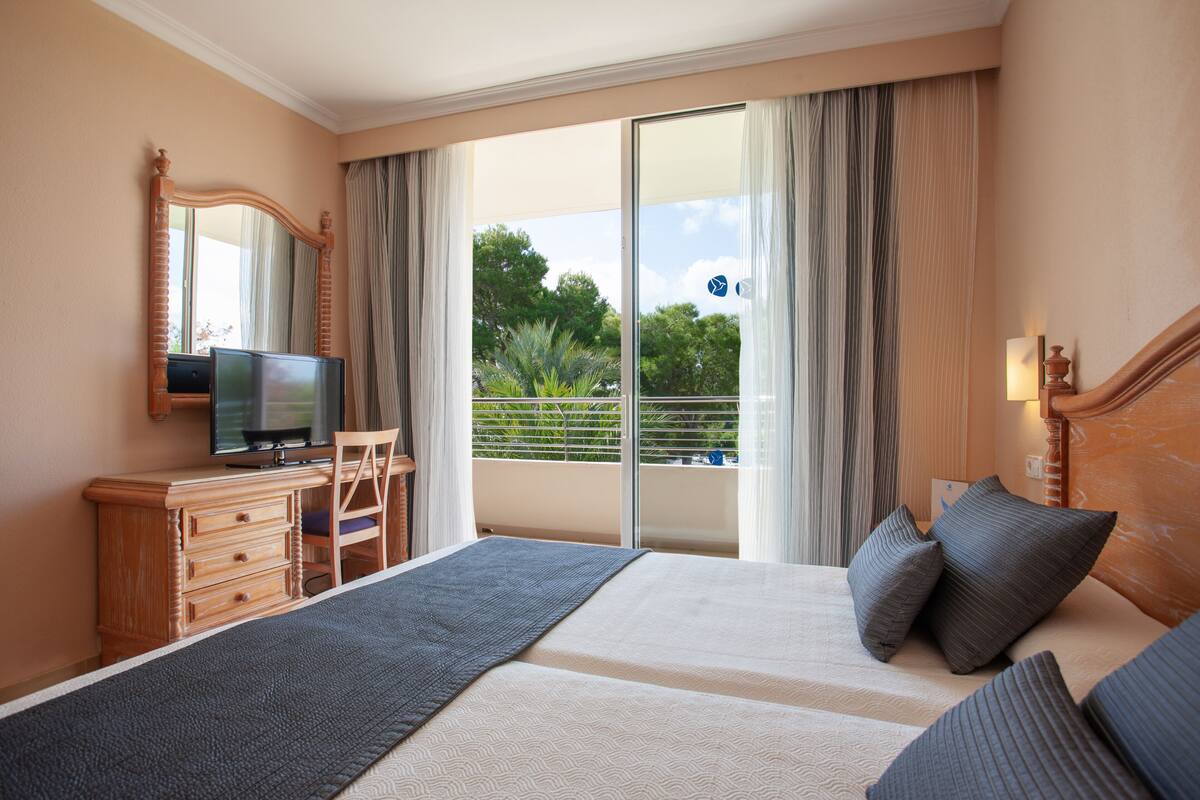 Ein Hotelzimmer mit einem Bett, einem Schreibtisch mit Fernseher, einem Sessel und einem Balkon mit Vorhängen.