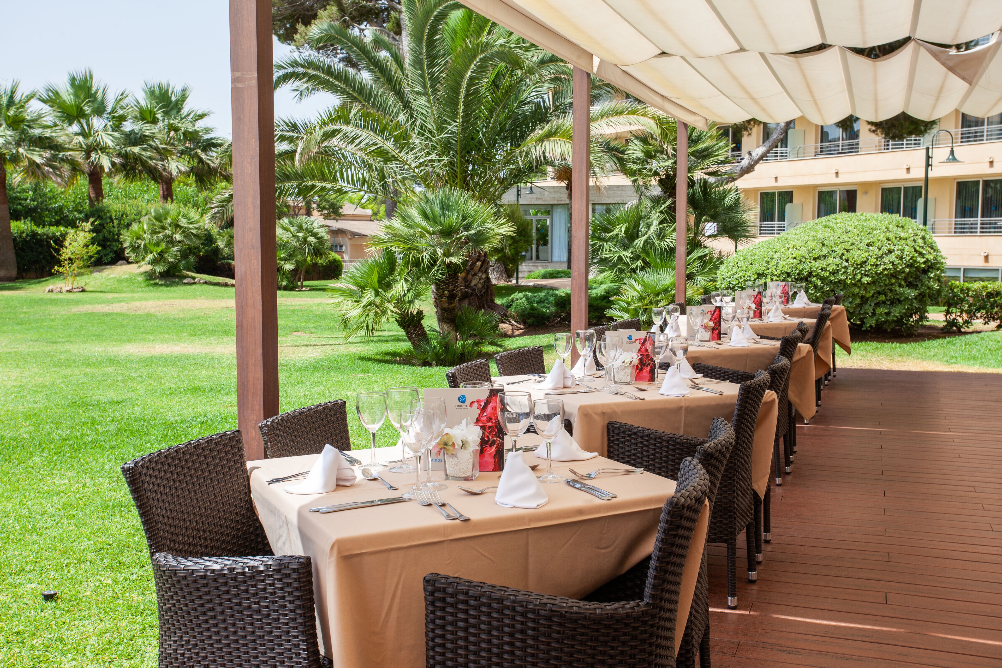 Un espace de restauration en plein air, avec des tables dressées pour un repas, entouré de verdure et avec un bâtiment en arrière-plan.