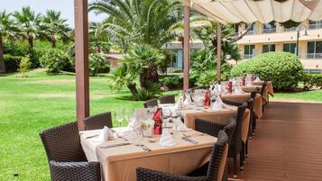 Un espace de restauration en plein air, avec des tables dressées pour un repas, entouré de verdure et avec un bâtiment en arrière-plan.