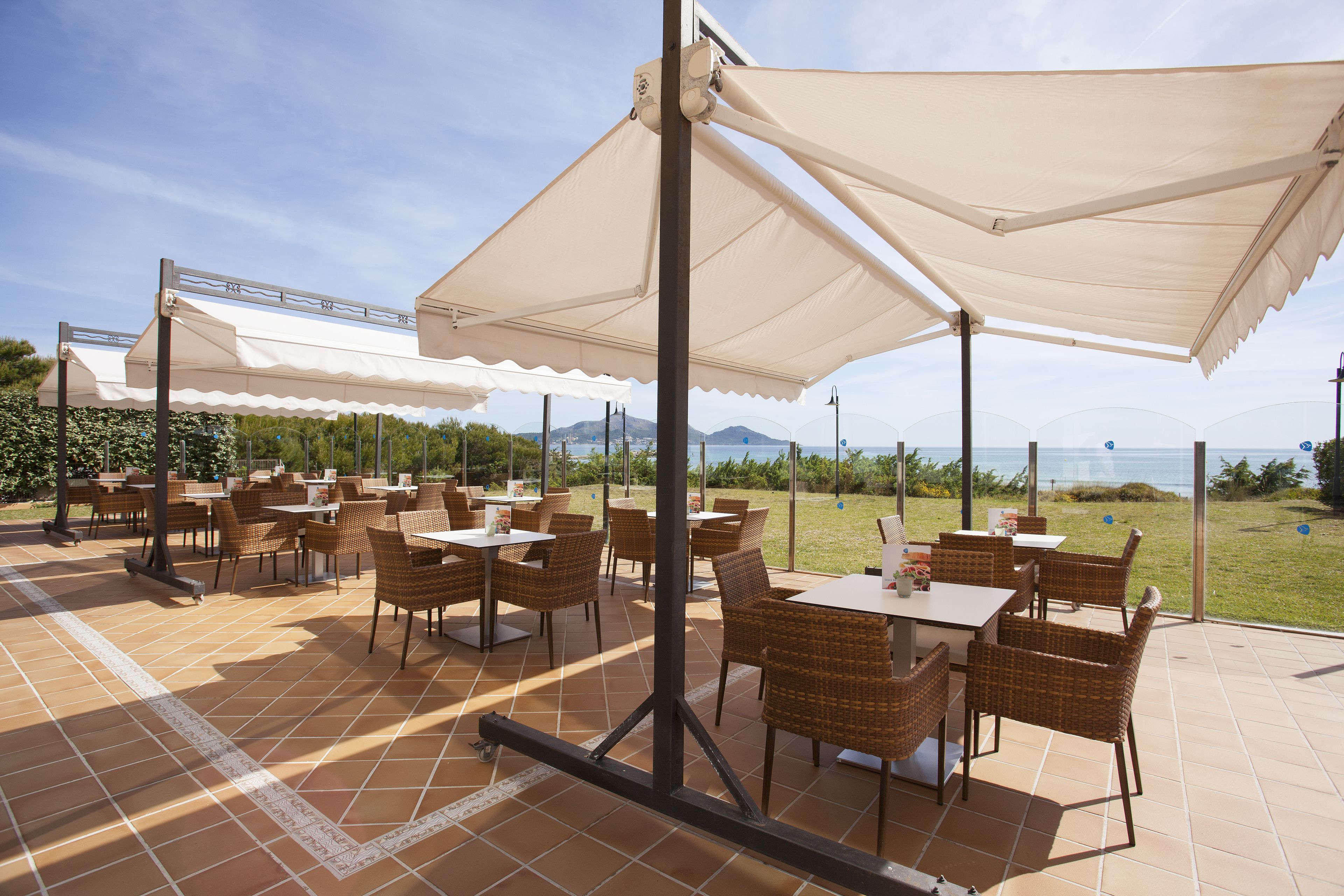 Een terras buiten met tafels en stoelen, grote parasols en uitzicht op zee.