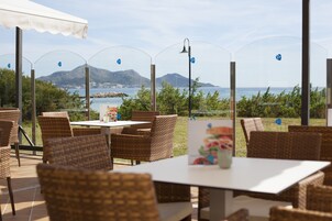 Un espacio de comedor al aire libre con sillas y mesas de mimbre, con vistas a un paisaje costero con montañas y el mar.