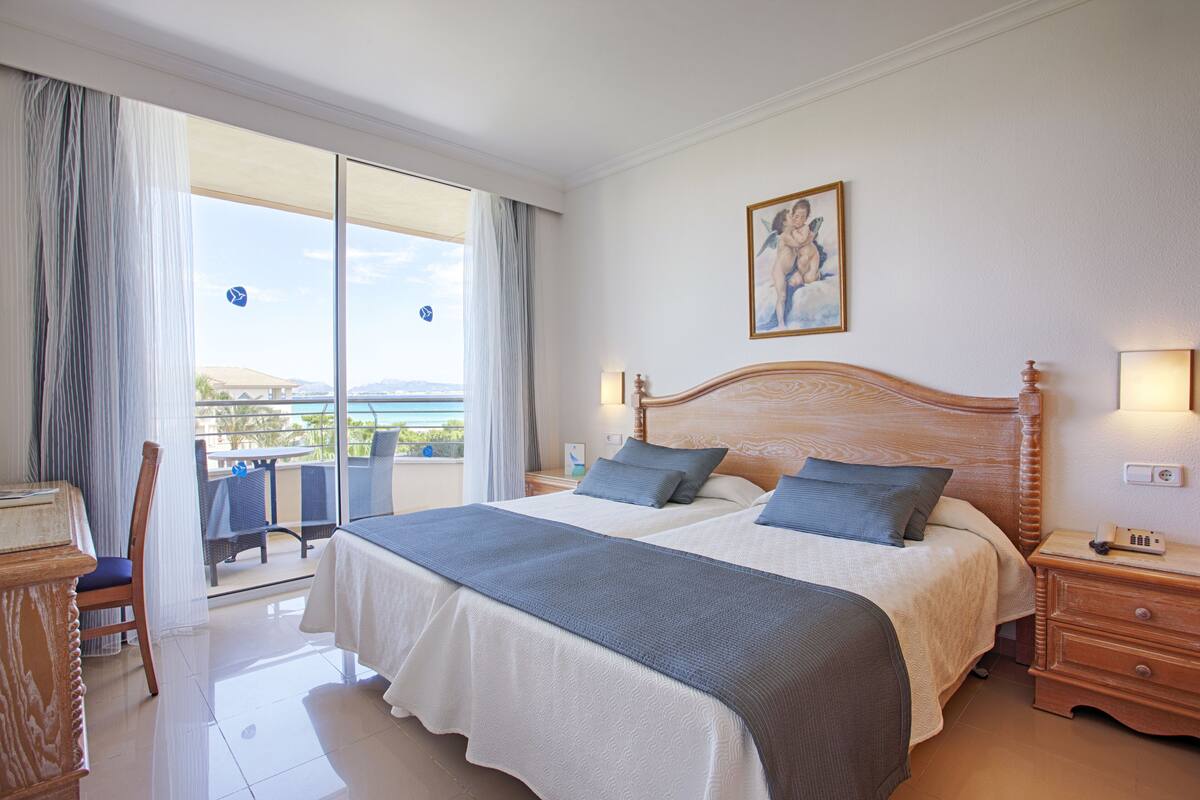 Ein Hotelzimmer mit einem großen Bett, einem hölzernen Kopfteil, einem Nachttisch, einem Schreibtisch und einem Stuhl. Ein Balkon bietet Blick auf den Ozean.