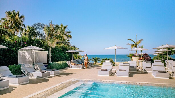 A poolside area with lounge chairs and umbrellas, a clear view of the ocean, and a tropical setting with palm trees. - Ivi Mare - Designed for adults by Louis Hotels (Geroskipou)