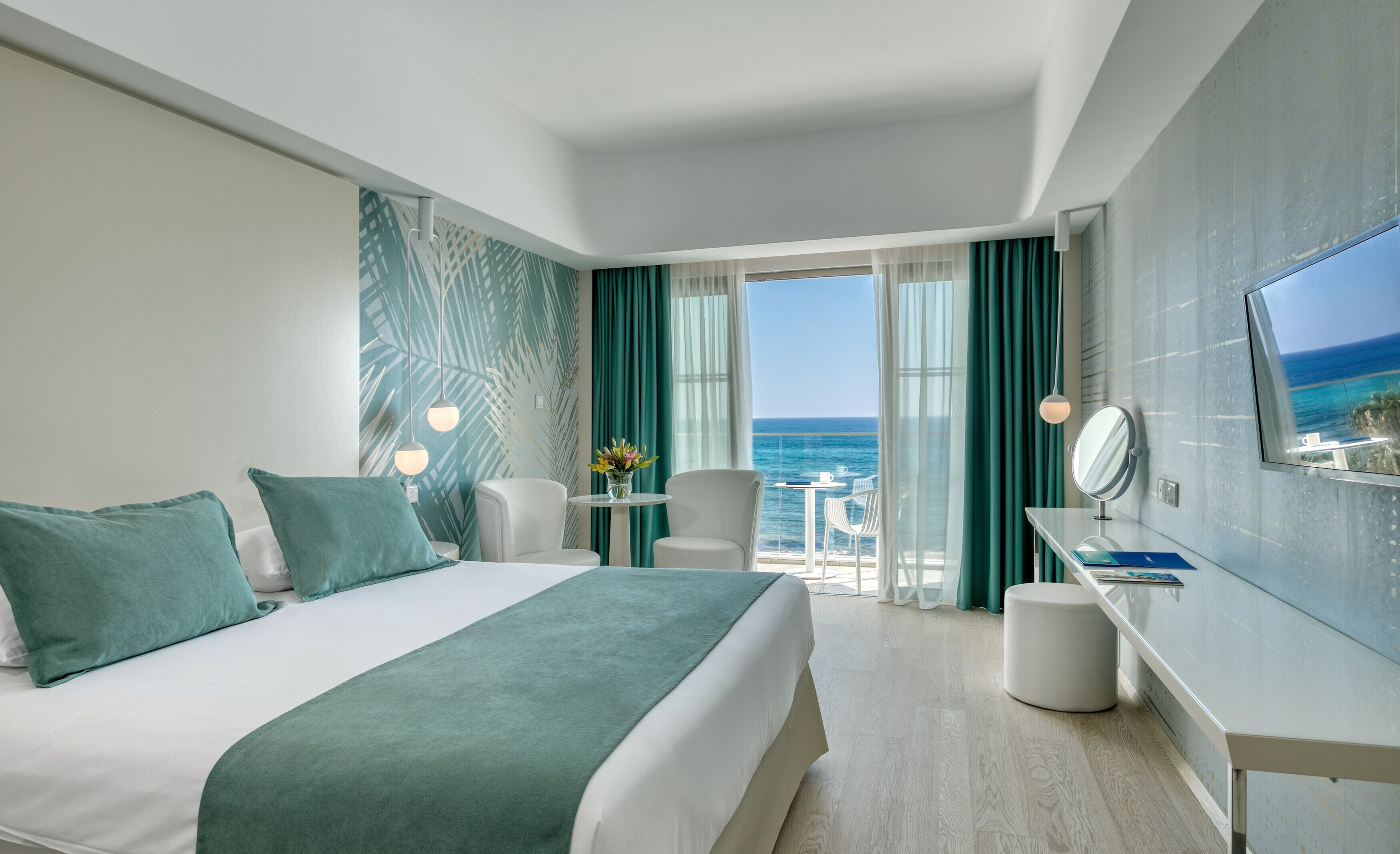 A hotel room with a bed, a desk, a chair, a TV, and a view of the ocean.