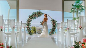 A wedding ceremony setup with a bride in a white dress, a floral arch, and white chairs arranged in rows. - Ivi Mare - Designed for adults by Louis Hotels (Geroskipou)
