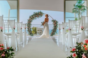 A wedding ceremony setup with a bride in a white dress, a floral arch, and white chairs arranged in rows. - Ivi Mare - Designed for adults by Louis Hotels (Geroskipou)
