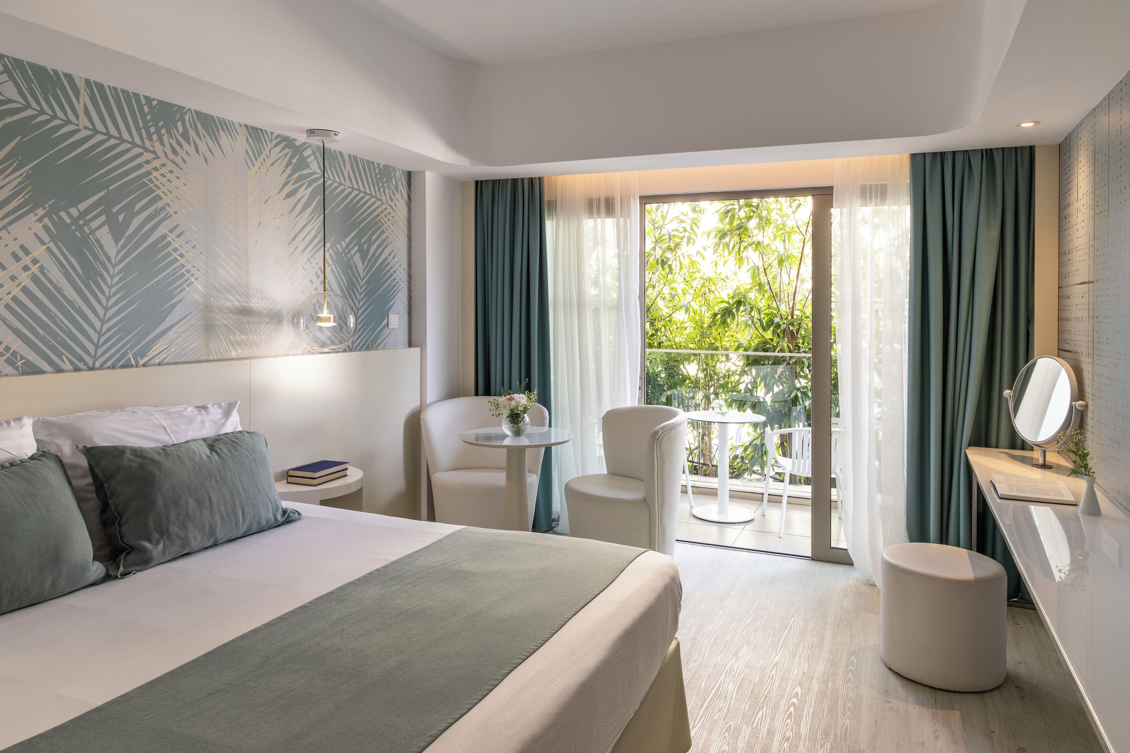 A modern hotel room with a large bed, a small round table, two chairs, a round mirror, and a balcony with a view of greenery.