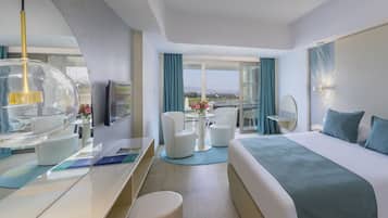 A modern hotel room with a large bed, a desk with a computer, a chair, a round table with flowers, and a view of the city.