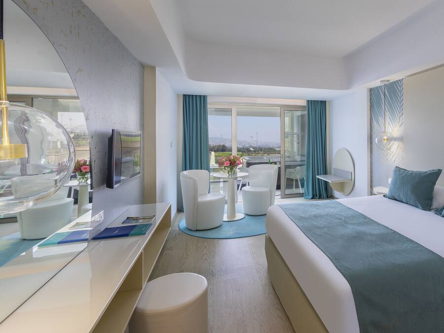 A modern hotel room with a large bed, a desk with a computer, a chair, a round table with flowers, and a view of the city.