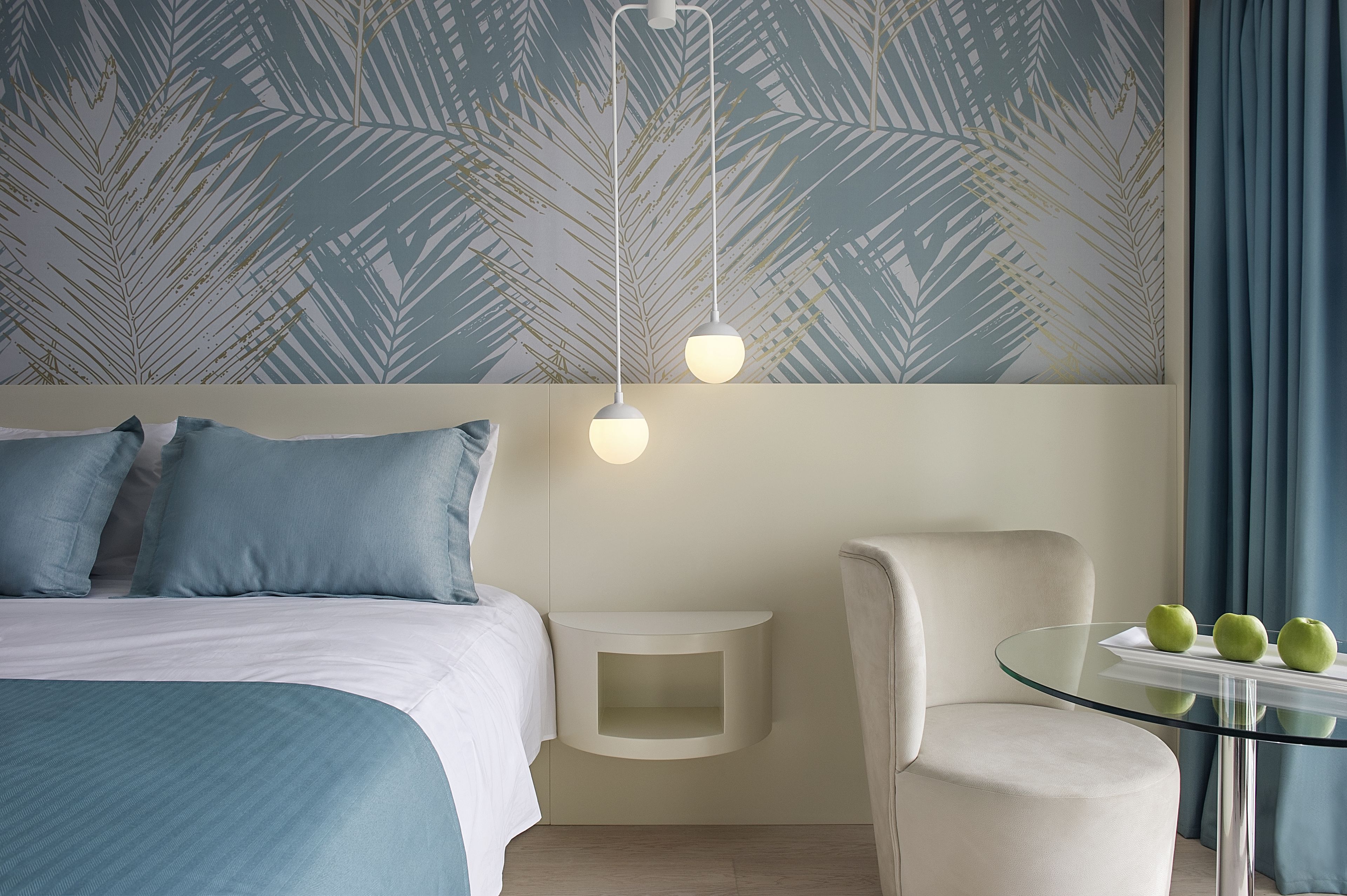 A modern hotel room with a bed, a side table, a glass table with apples, and a chair.