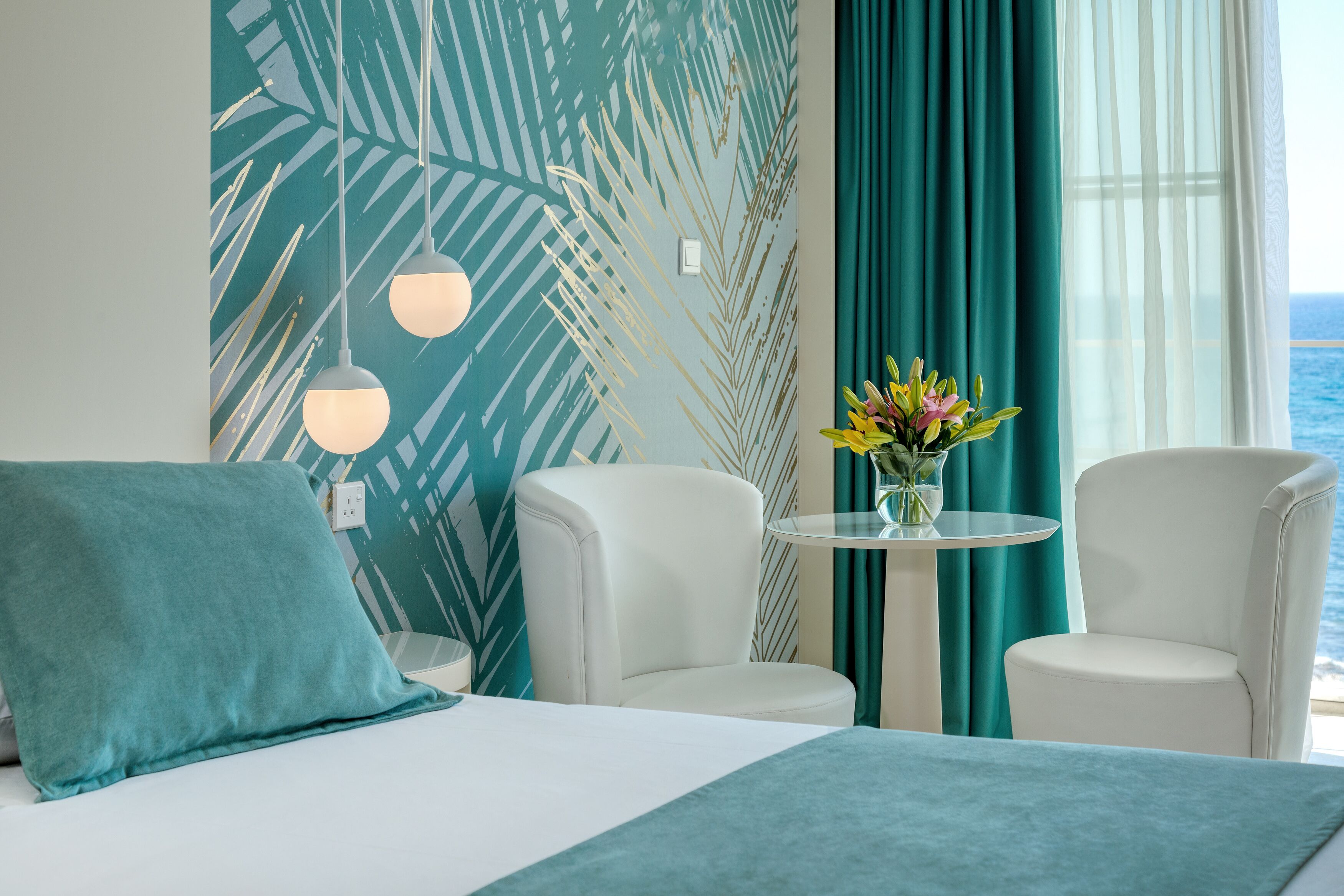 A hotel room with a bed, two white armchairs, a small round table with a vase of flowers, teal curtains, and a view of the sea.