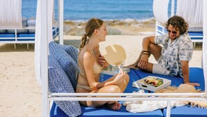 A beachside lounge with a view of the ocean, a dining table with food, and two people relaxing on a blue lounge chair. - Ivi Mare - Designed for adults by Louis Hotels (Geroskipou)