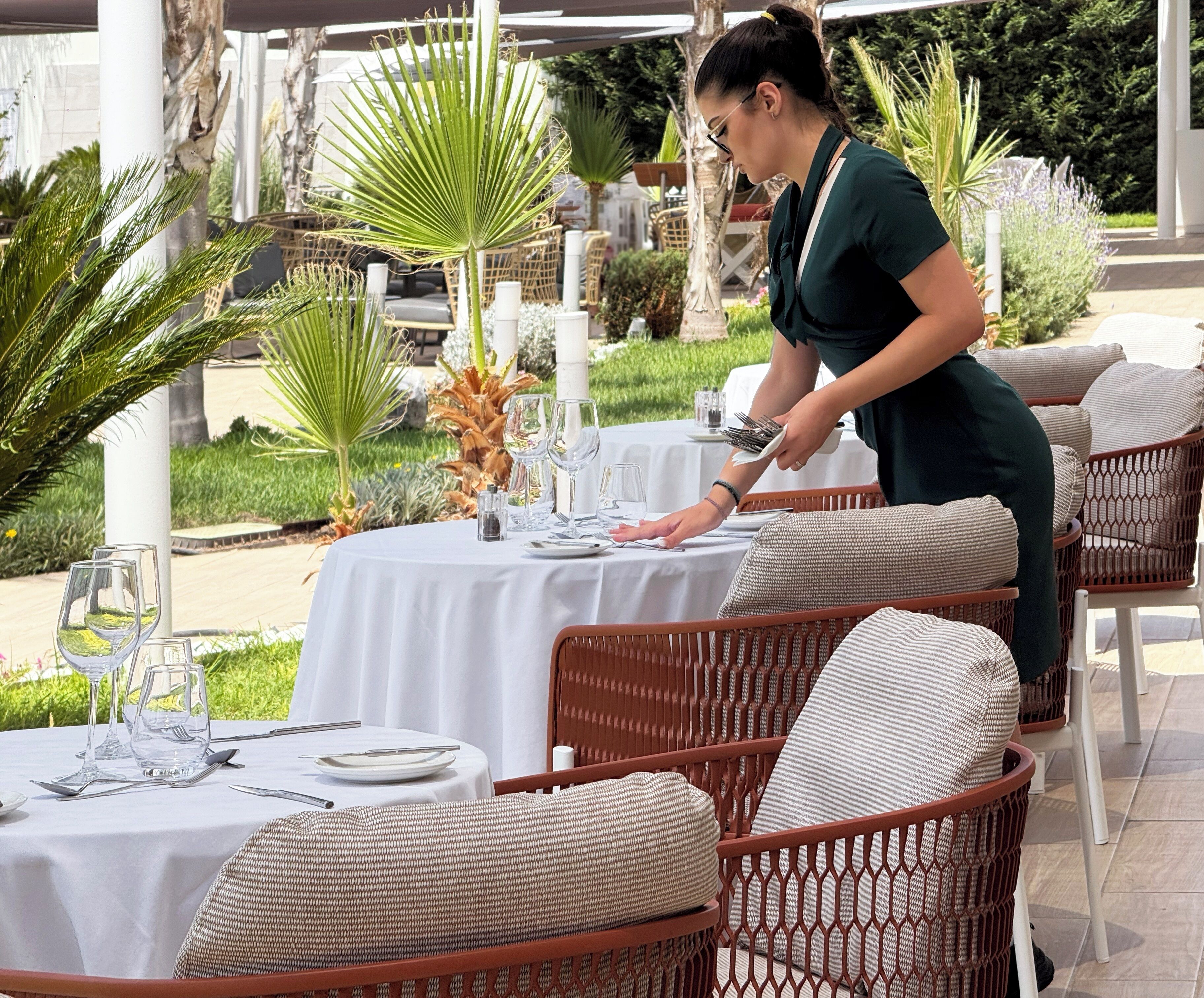Comedor con sillas de mimbre y manteles blancos, una persona sirviendo y vista a un jardín.