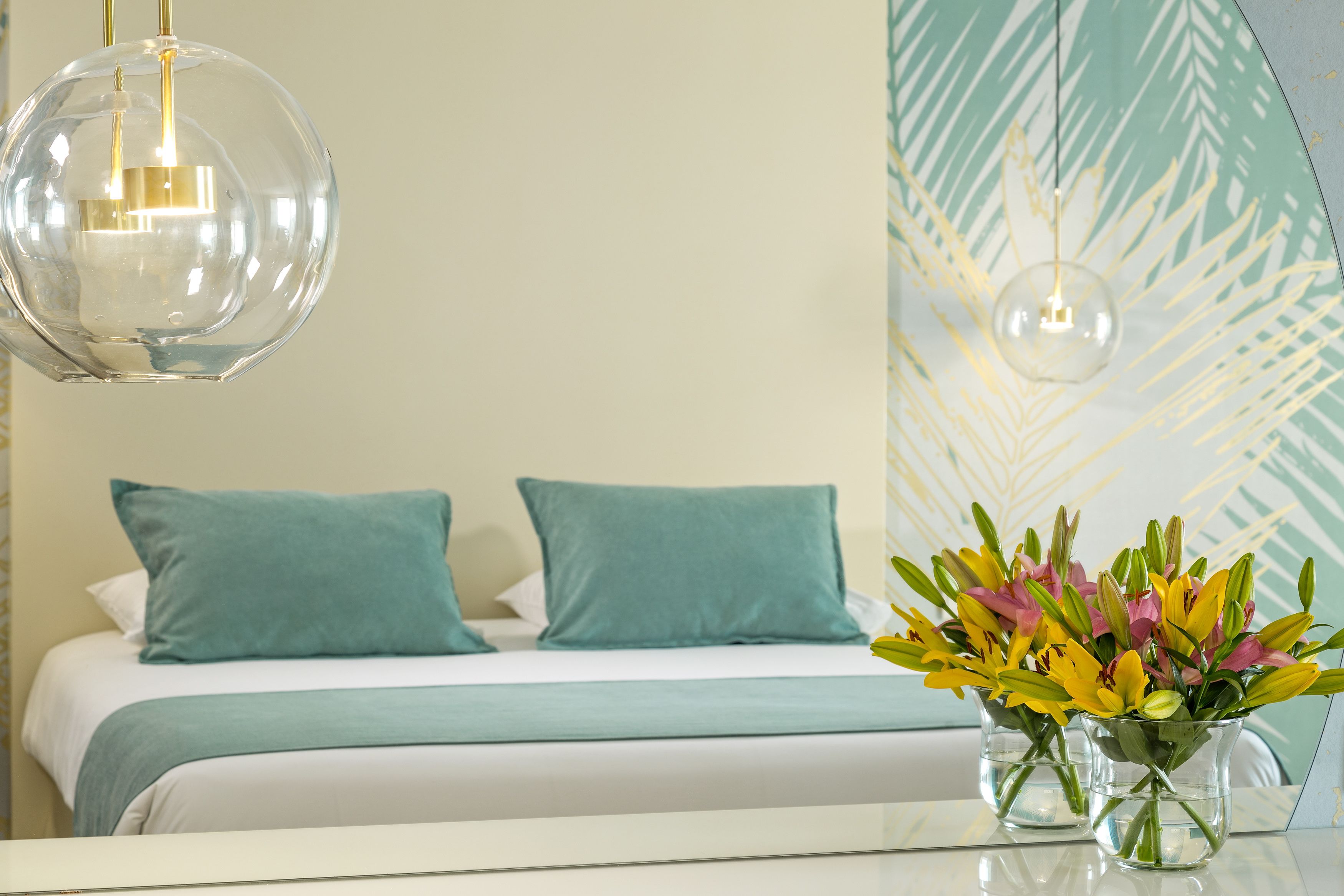 Habitación de hotel con cama, almohadones azul turquesa, lámpara colgante de cristal transparente y un jarrón con flores.