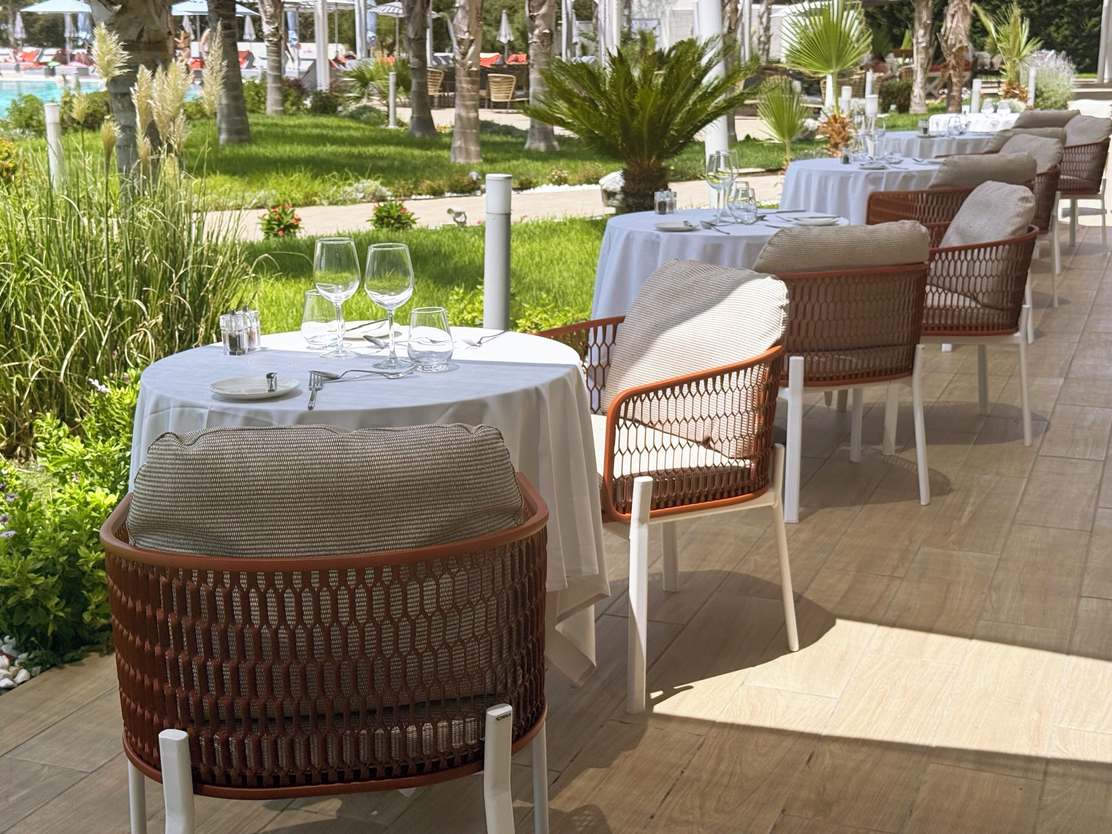 Eine Terrasse mit runden Esstischen, Korbstühlen und einem Pool im Hintergrund.
