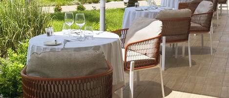 A terrace with round tables set for dining, wicker chairs, and a pool in the background.