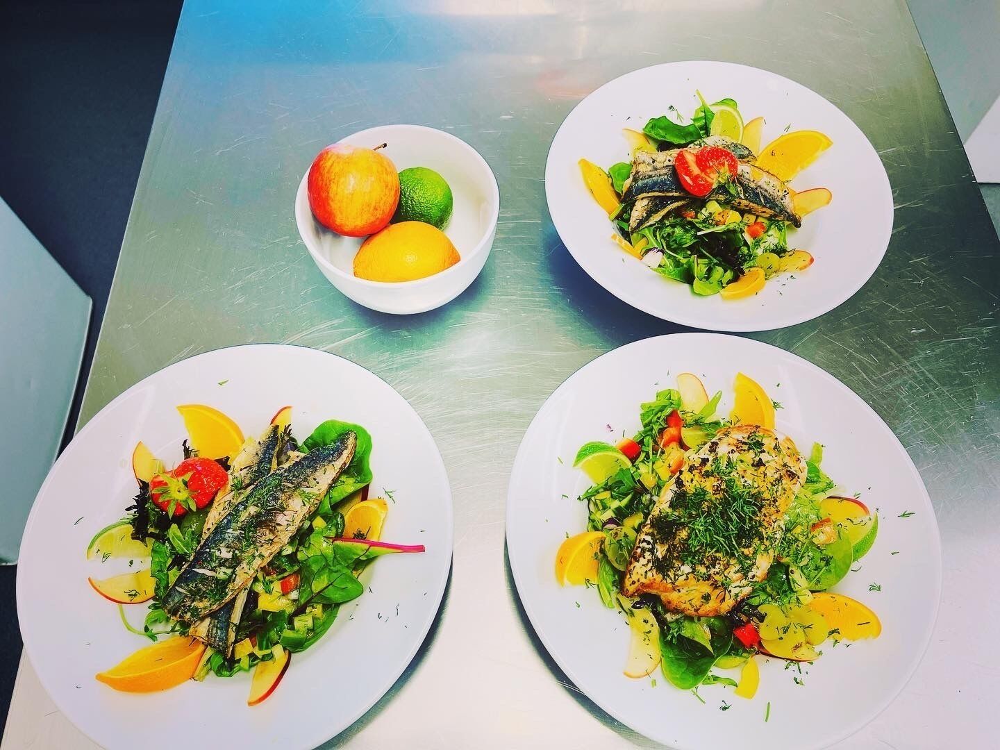 Tres platos de comida con pescado y verduras sobre una mesa de acero inoxidable.