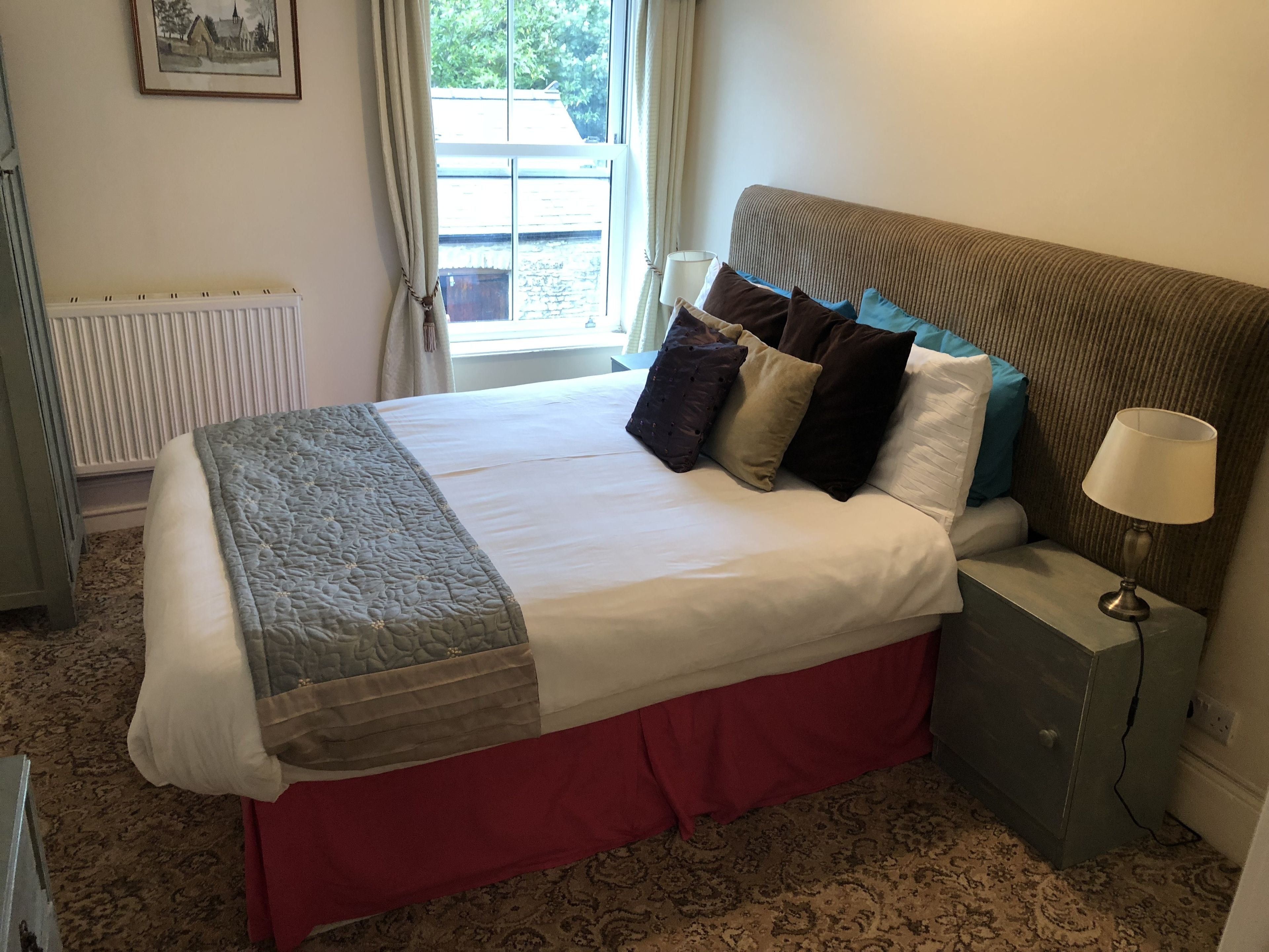 A hotel room with a large bed, a bedside table with a lamp, a radiator, and a window with curtains.