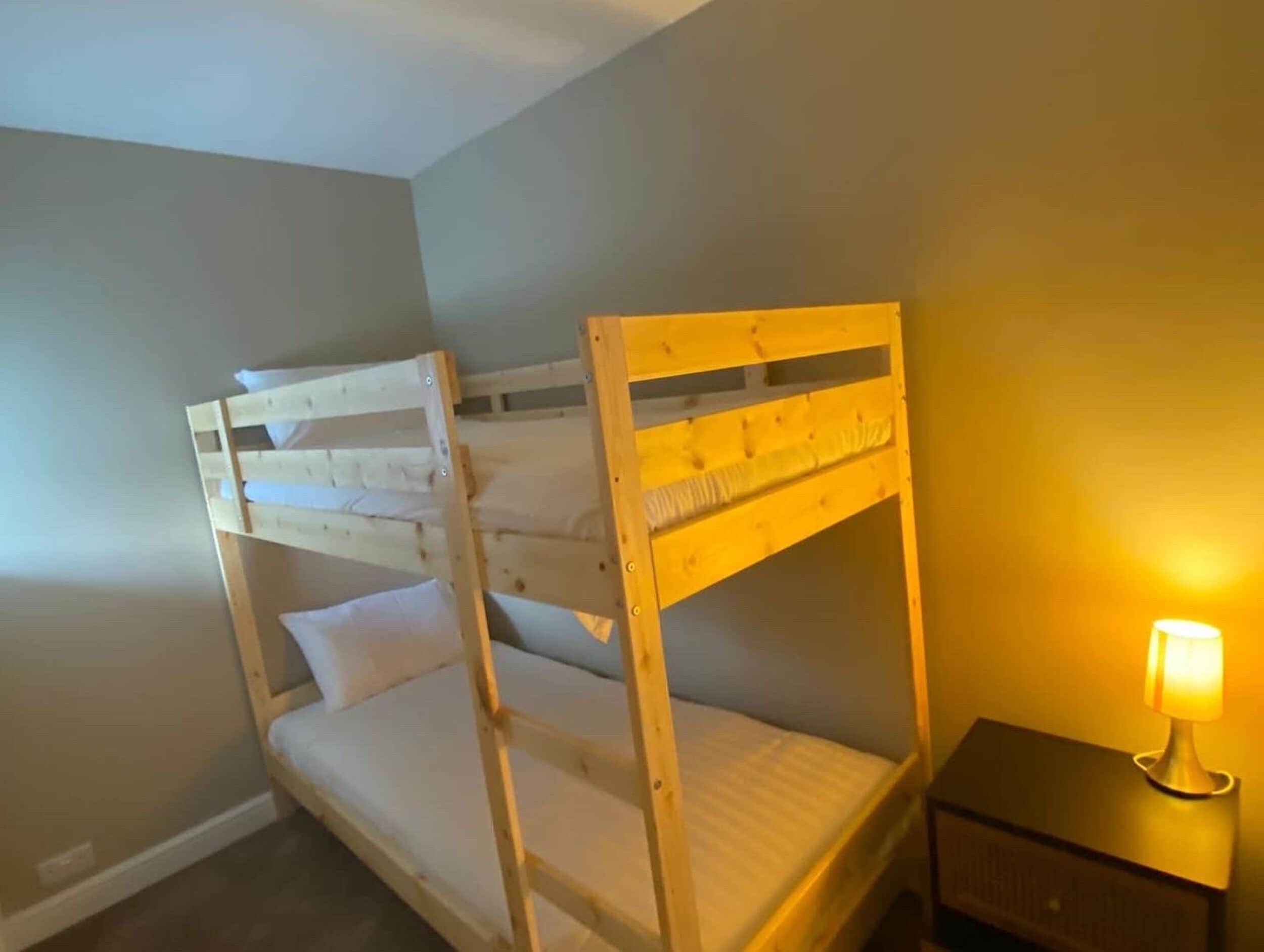 A bunk bed with a wooden frame and white bedding, a bedside table with a lamp, and a plain wall.