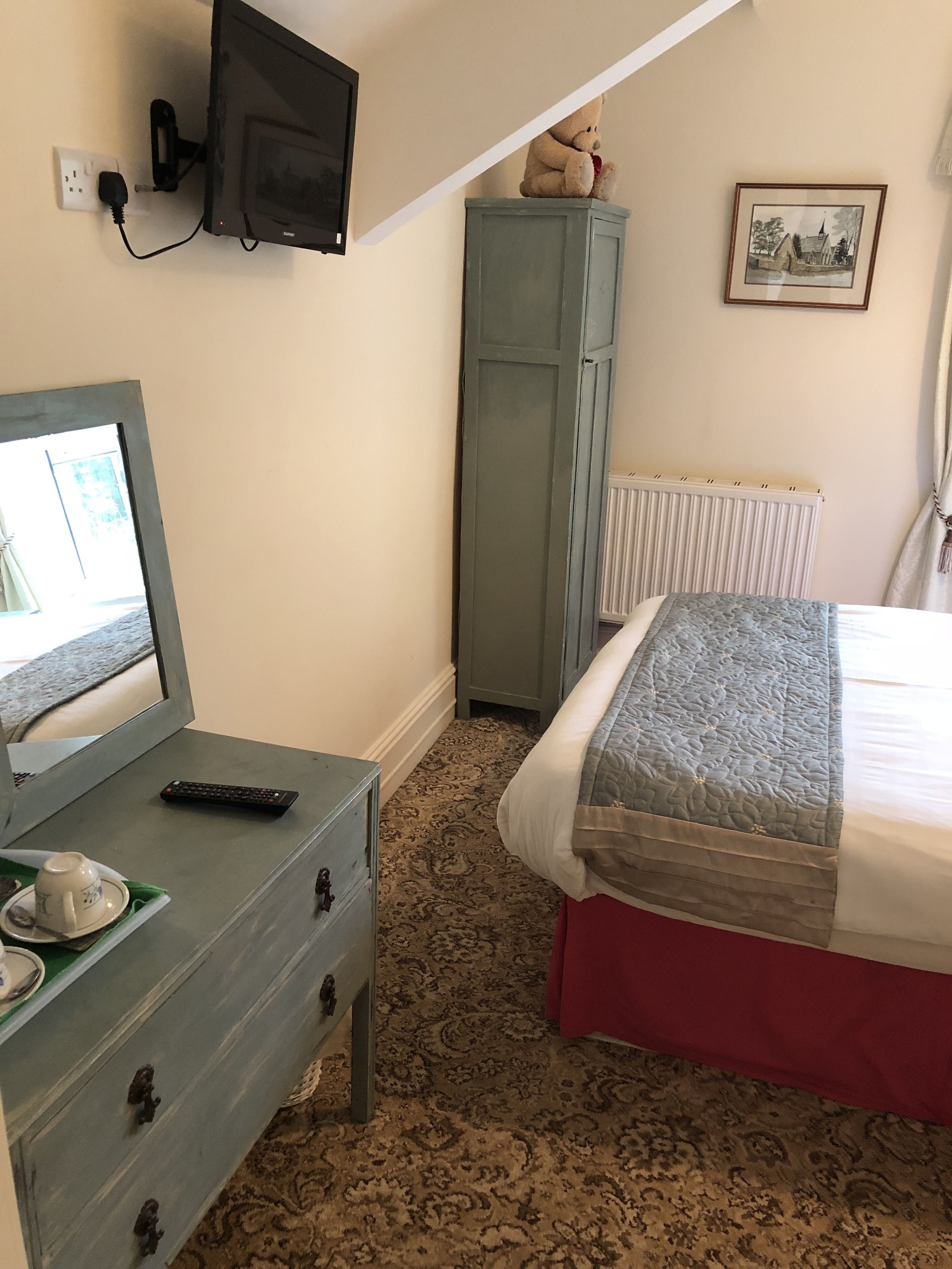 A bedroom with a bed, a dresser, a television mounted on the wall, a radiator, and a teddy bear on a cabinet.