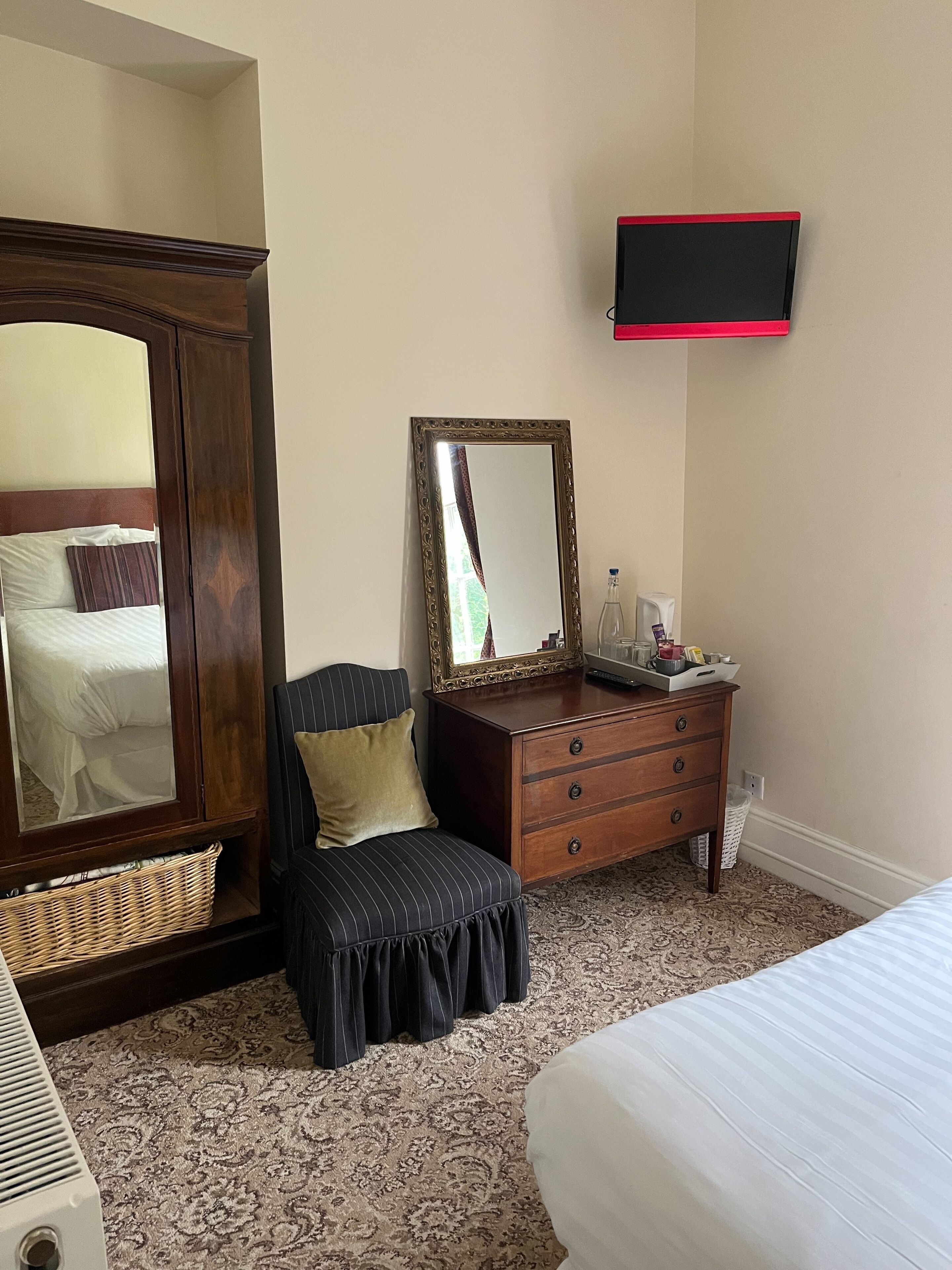A hotel room with a large mirror, a wooden wardrobe, a bedside table with a lamp, a small stool, and a bed.