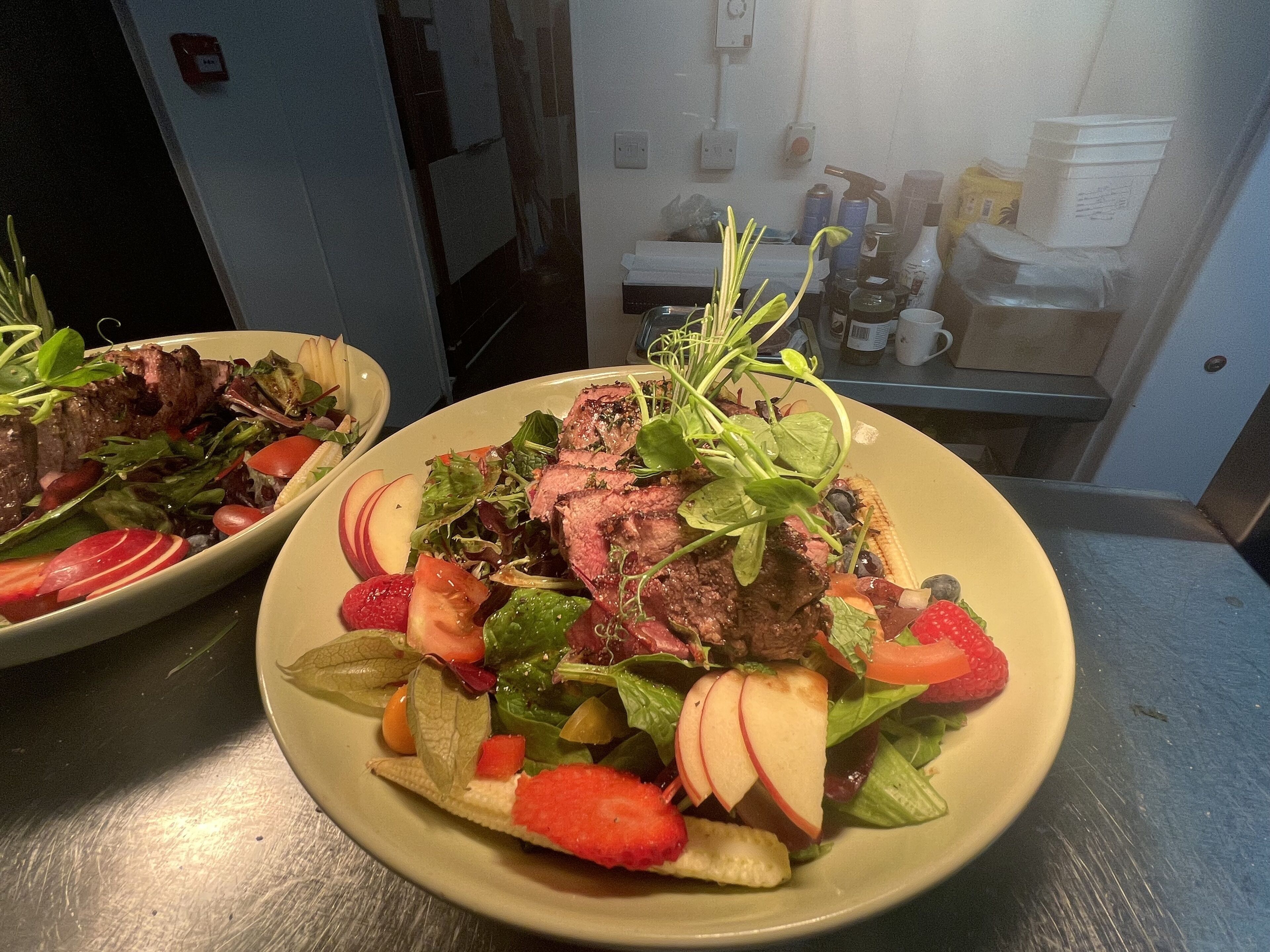 Two plates of food with steak and salad on a stainless steel counter.