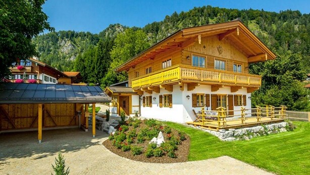 A two-story wooden chalet with a balcony, surrounded by a well-maintained garden and a paved driveway. - Chalet unter Weißblauem Himmel (Reit im Winkl)