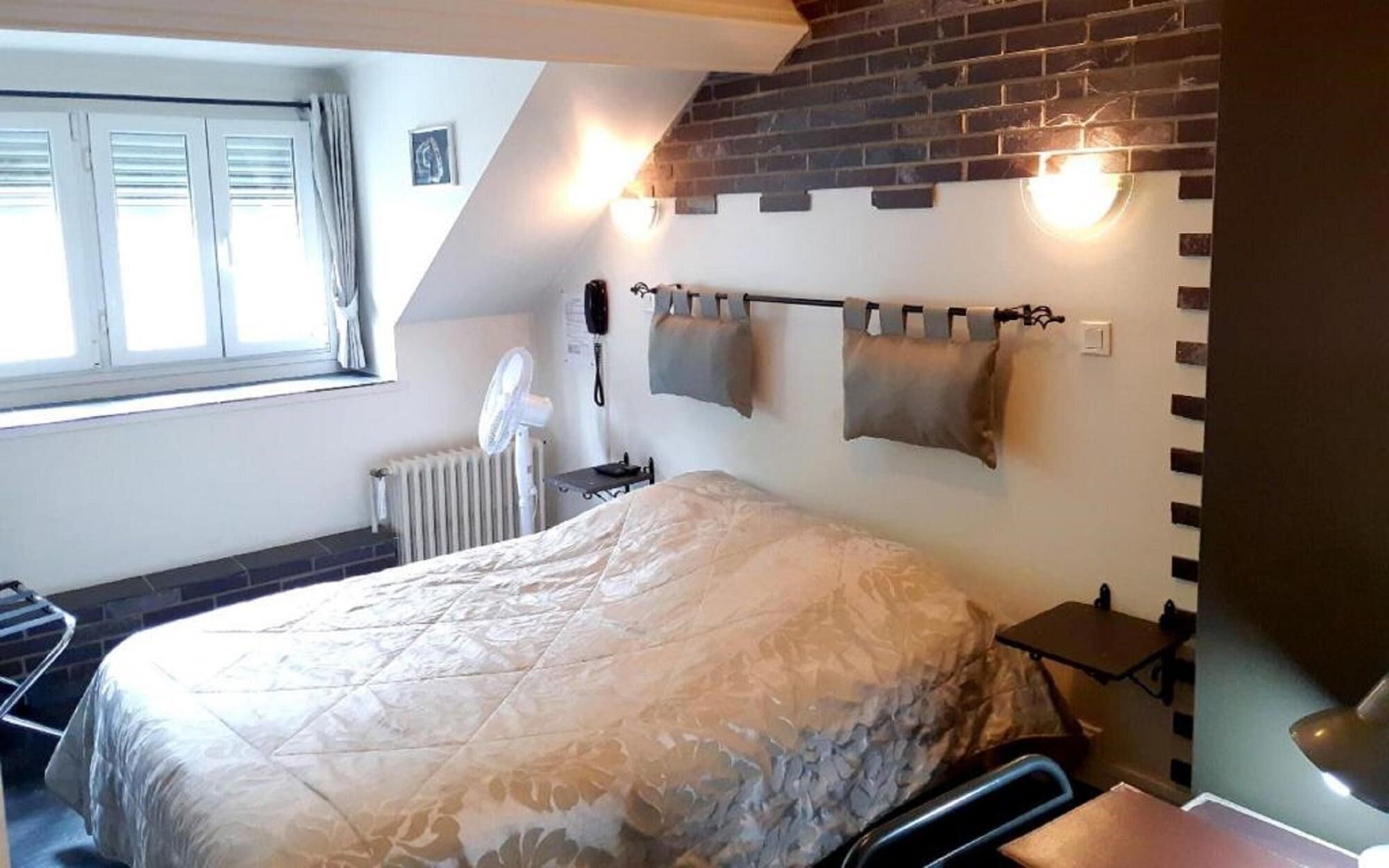 A bedroom with a bed, a radiator, a window with curtains, a wall-mounted light, and a brick wall.