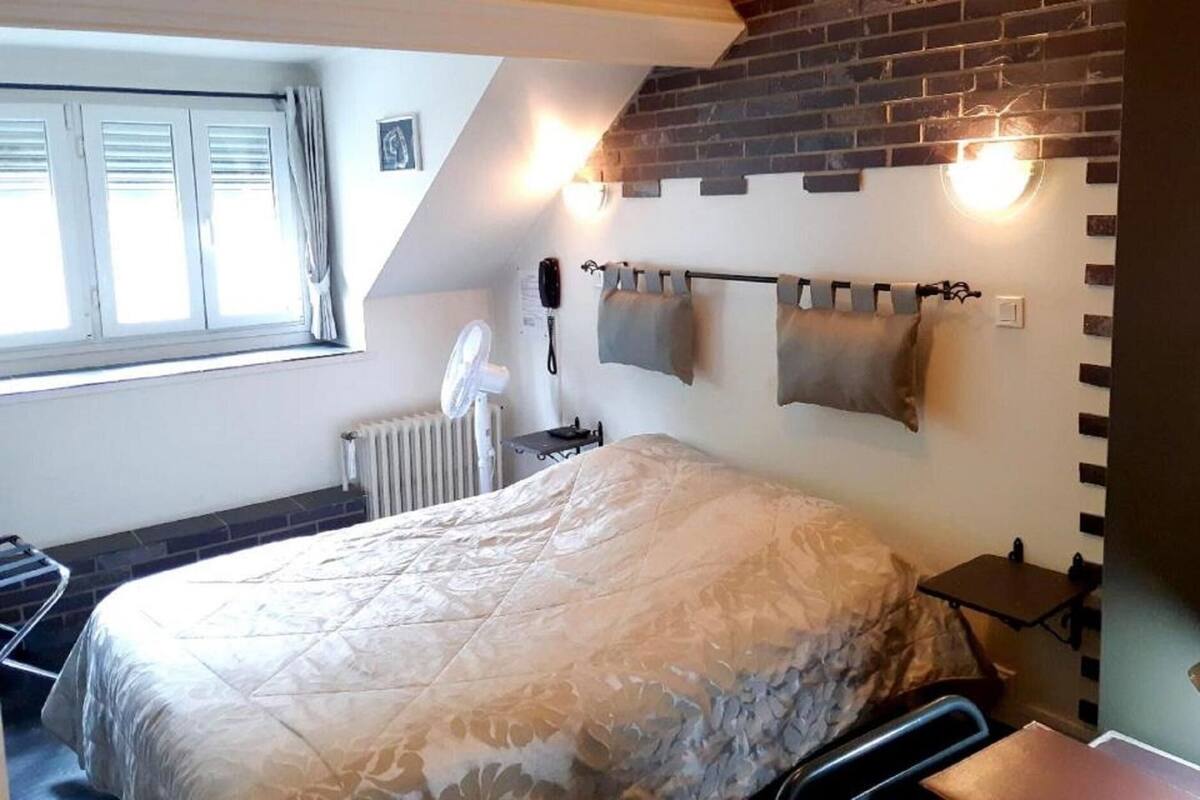 A bedroom with a bed, a radiator, a window with curtains, a wall-mounted light, and a brick wall.