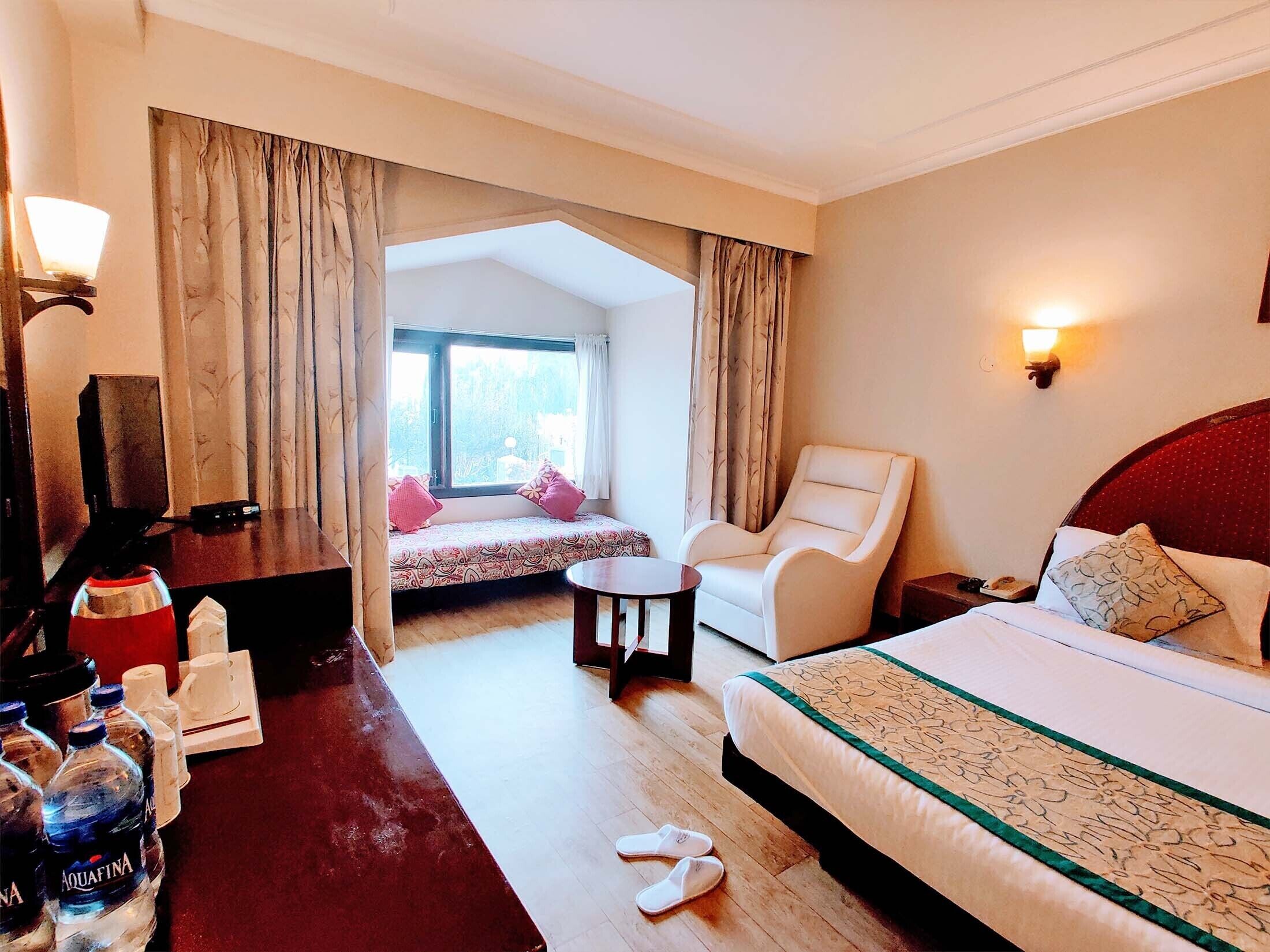 A hotel room with a bed, a chair, a desk with a lamp, a window with curtains, and a view of the outdoors.