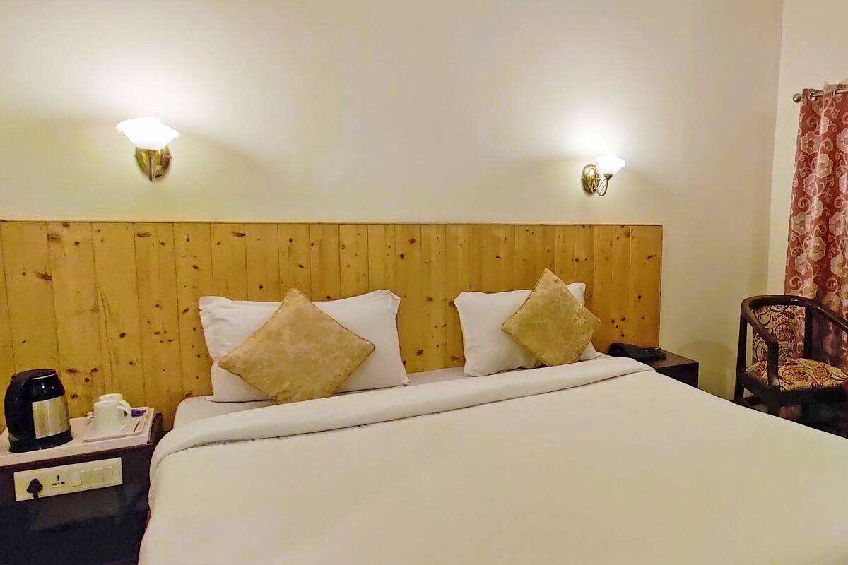 A hotel room with a wooden headboard, a bed with white linens, two wall-mounted lights, a nightstand with a coffee maker, and a chair with a floral cushion.