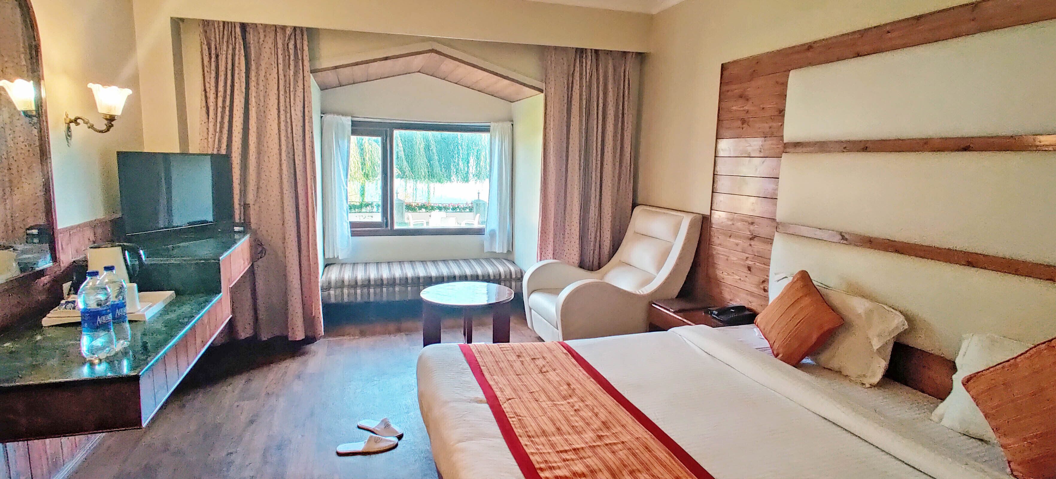 A hotel room with a bed, a desk with a TV, a chair, a small table, and a window with curtains.