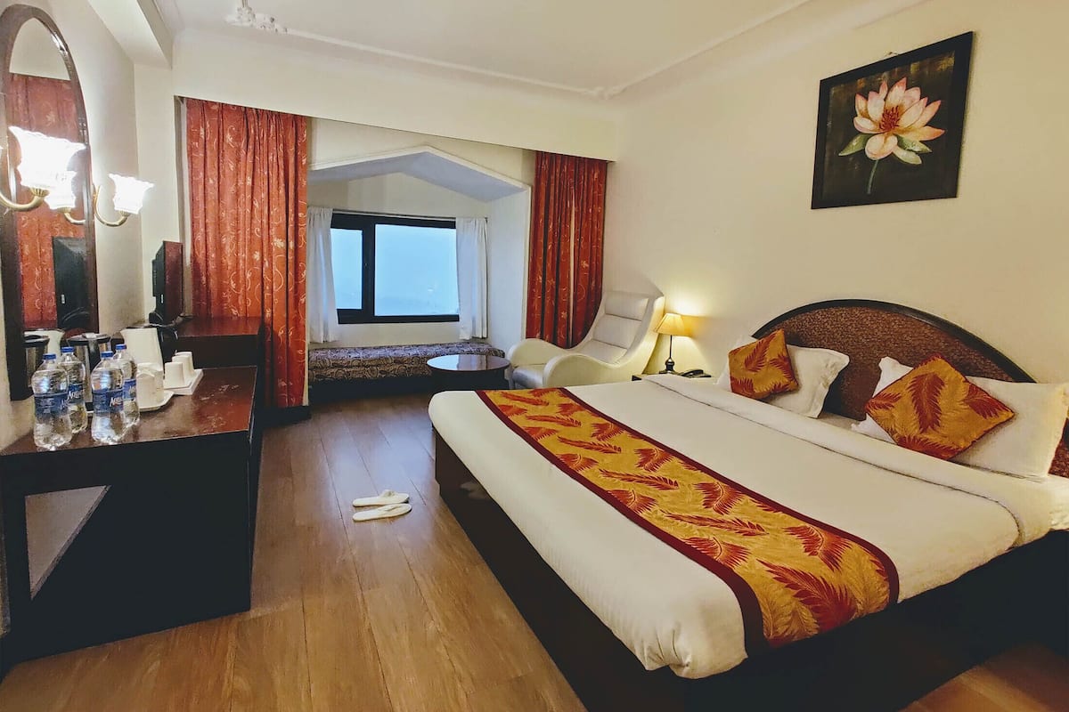 A hotel room with a bed, a desk with a mirror, a chair, a window with curtains, and a painting on the wall.