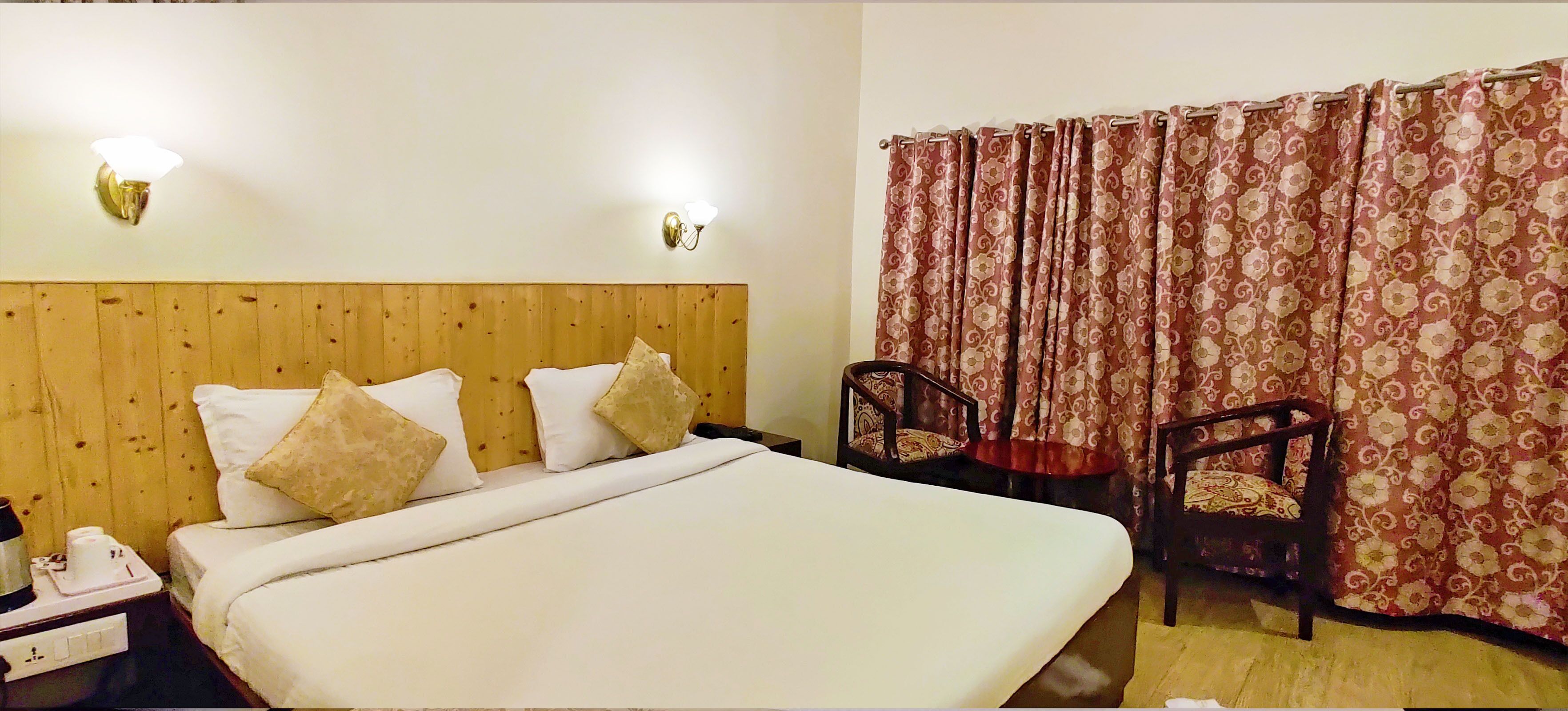 A hotel room with a wooden headboard, a large bed with white linens, two wall-mounted lights, a pair of chairs, and a curtain with a floral pattern.