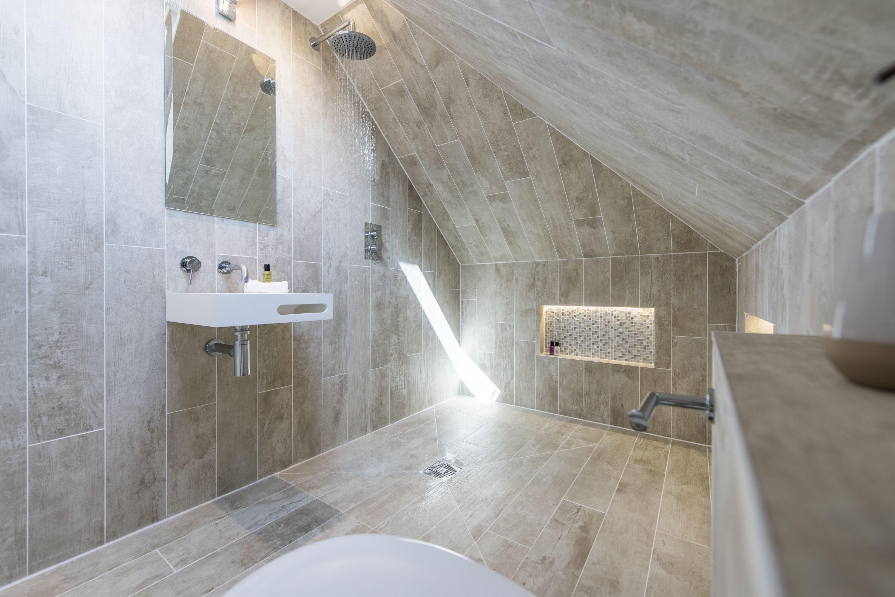 A modern bathroom with a sloped ceiling, a built-in sink, a shower, and a large mirror.