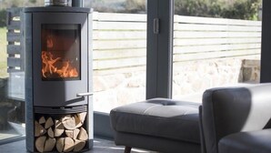 A modern fireplace with a glass door, a stack of firewood inside, and a view of a stone wall and greenery outside. - Chyreen@marazion (Marazion)