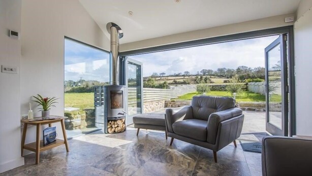 A modern living room with a stone-tiled floor, a grey sofa, a small table with a plant, and a wood-burning stove. - Chyreen@marazion (Marazion)