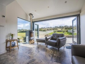 A modern living room with a stone-tiled floor, a grey sofa, a small table with a plant, and a wood-burning stove. - Chyreen@marazion (Marazion)