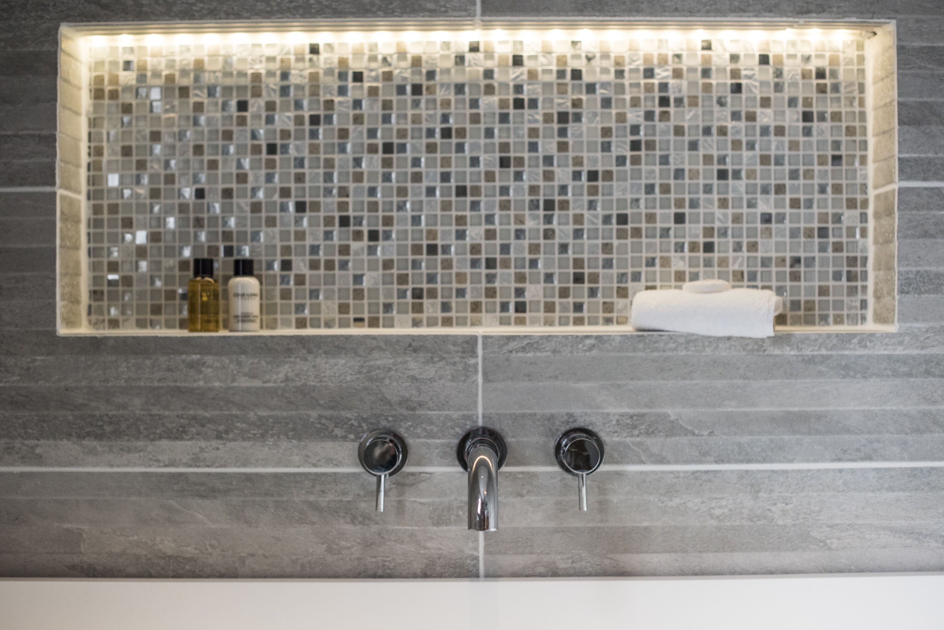 A bathroom with a tiled mosaic backsplash, a sink with three faucets, and a towel rack.
