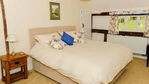 Een slaapkamer met een groot bed, een nachtkastje met een lamp, een lijst met een afbeelding aan de muur en een raam met bloemen gordijnen.