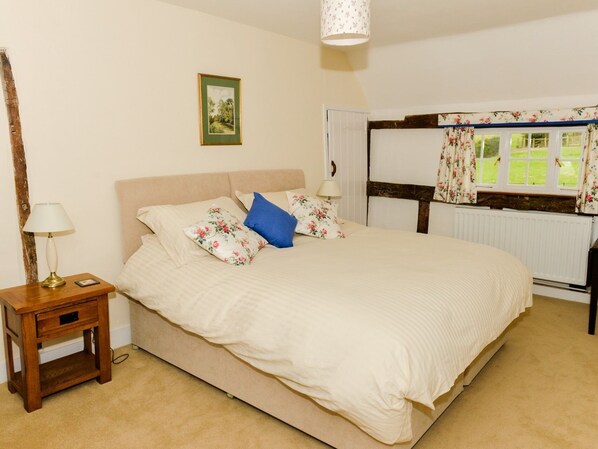 A bedroom with a large bed, a nightstand with a lamp, a framed picture on the wall, and a window with floral curtains.