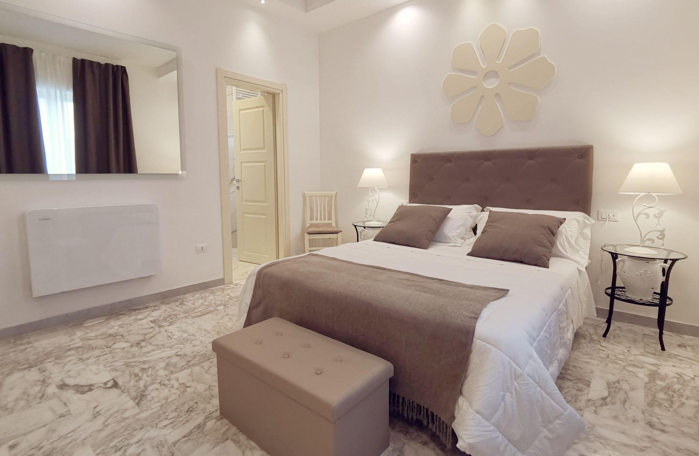 A bedroom with a large bed, two bedside tables with lamps, a bench, a mirror, and a door.