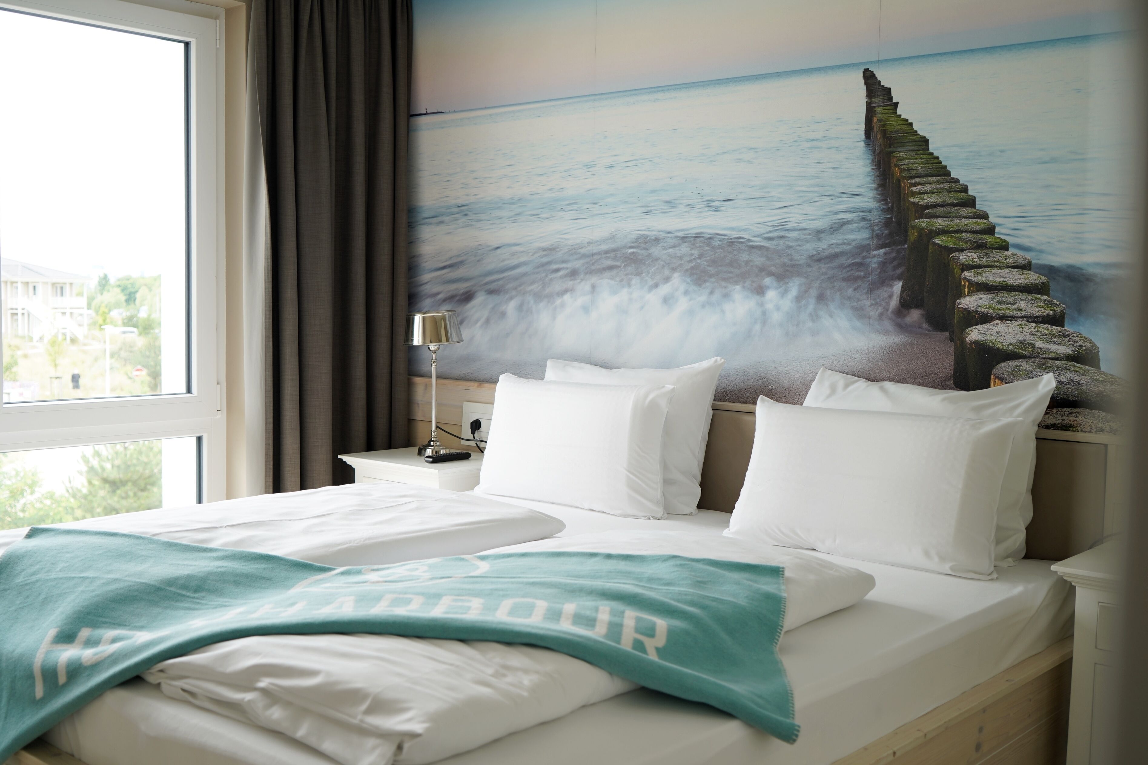 Una habitación de hotel con cama, ropa de cama blanca, un plaid verde azulado, una mesita de noche con lámpara y un mural de un muelle que se extiende hacia el mar.