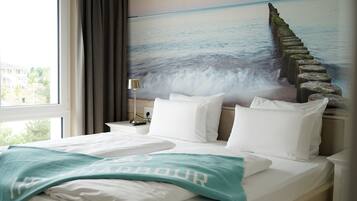 Una habitación de hotel con cama, ropa de cama blanca, un plaid verde azulado, una mesita de noche con lámpara y un mural de un muelle que se extiende hacia el mar.