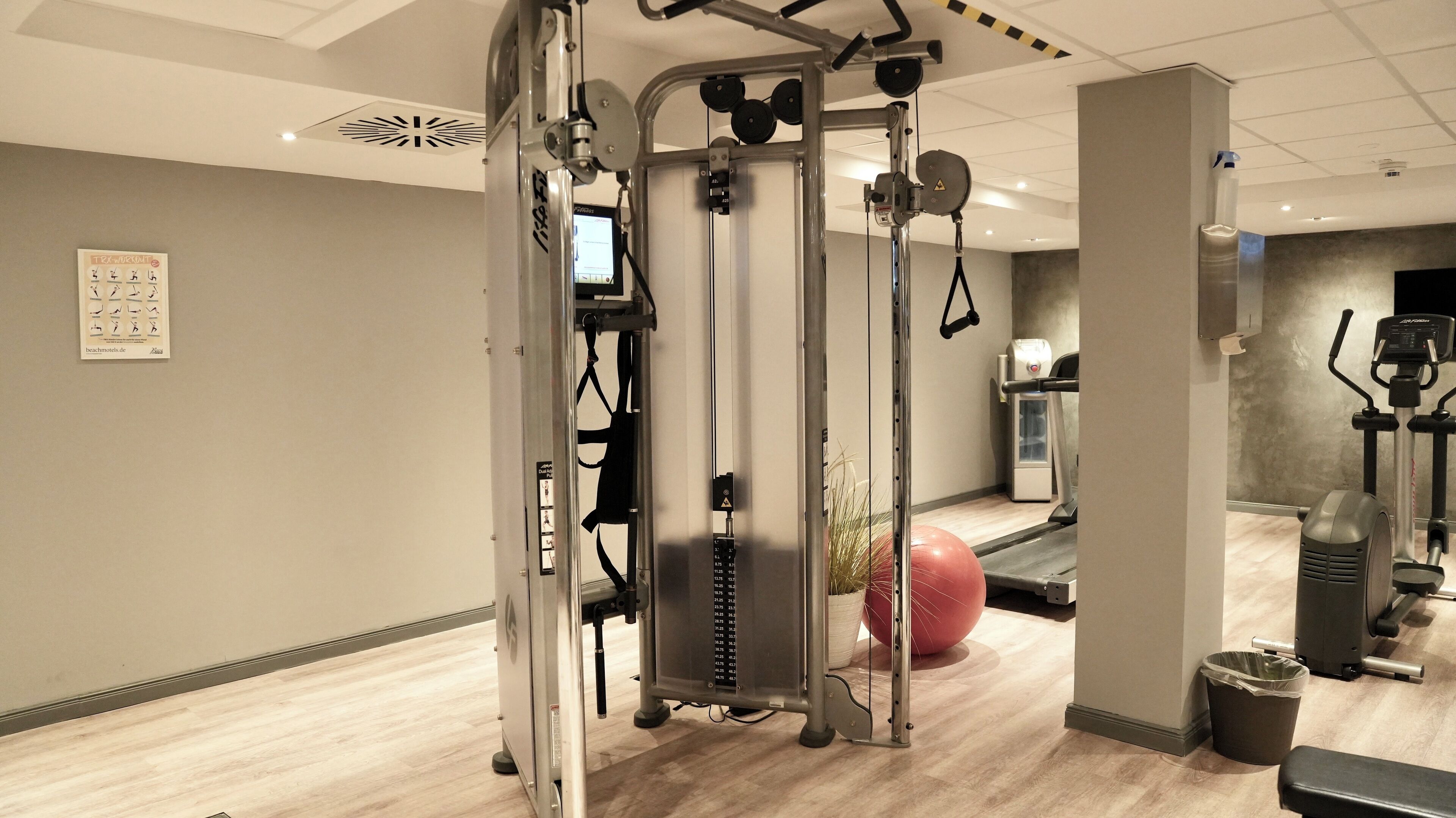 Een fitnessruimte met diverse trainingsapparaten, waaronder een kabeltrekapparaat, loopband en hometrainer.