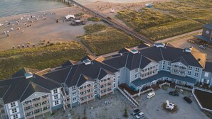 Ovenfra set et kysthotel med flere bygninger, en mole der strækker sig ud i vandet, og en strand med strandstole og parasoller.