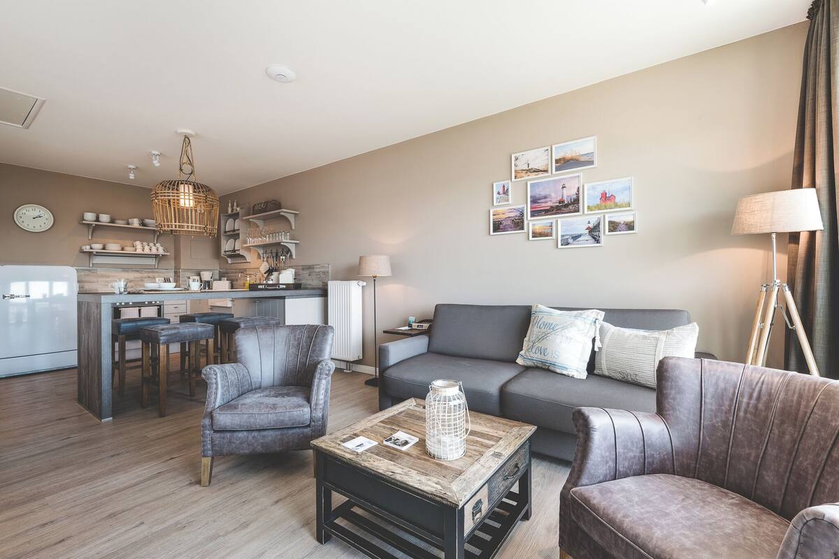 Un salon moderne avec un canapé gris, deux fauteuils, une table basse et un coin cuisine en arrière-plan.